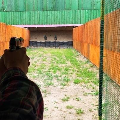 Strzelnica: osoba celuje z pistoletu w tarczę, widoczne ogrodzenie z siatki, zielone i pomarańczowe drewniane ściany boczne.