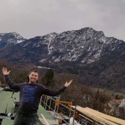 Uśmiechnięty mężczyzna na dachu z zieloną membraną dachową na tle górskim, z widocznymi rusztowaniami i elementami konstrukcyjnymi.