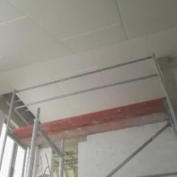 Montaż płyt kartonowo-gipsowych na suficie, widoczne rusztowanie i fragment ściany z bloków, ocieplenie wełną mineralną widoczne w otworze.