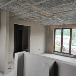 Surowe wnętrze po tynkowaniu maszynowym: ściany pokryte szarym tynkiem, widoczna surowa betonowa konstrukcja stropu, niezabezpieczone okno z widokiem na zewnątrz.