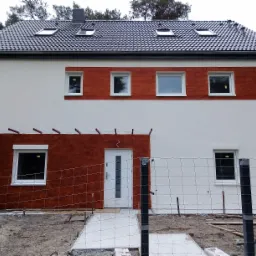 Dwukondygnacyjny dom jednorodzinny z jasną elewacją częściowo pokrytą imitacją drewna, widok z zewnątrz, teren wokół domu w trakcie prac wykończeniowych, ogrodzenie z siatki.