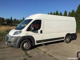 Biały furgon dostawczy Fiat Ducato z wysokim dachem, zaparkowany na betonowym placu z zielenią w tle, widoczny z boku w słoneczny dzień.