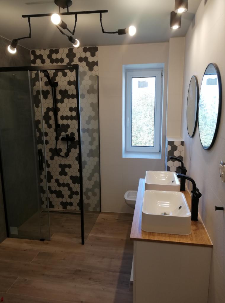 Nowoczesna łazienka po remoncie: kabina prysznicowa z wzorem heksagonalnym, dwie umywalki na drewnianym blacie, okrągłe lustra i stylowe oświetlenie.