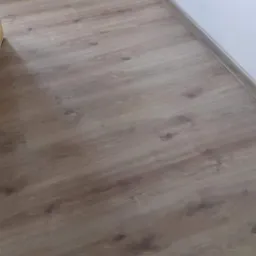 Jasne panele podłogowe z widoczną strukturą drewna, ułożone wzdłuż białej ściany z gniazdkiem elektrycznym.