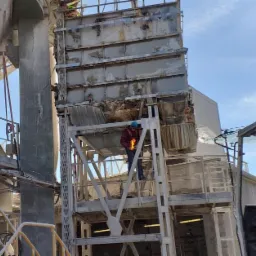 Pracownicy w kaskach i odblaskowych kamizelkach pracują przy demontażu lub naprawie konstrukcji stalowej na terenie przemysłowym, widoczny dźwig i elementy instalacji.