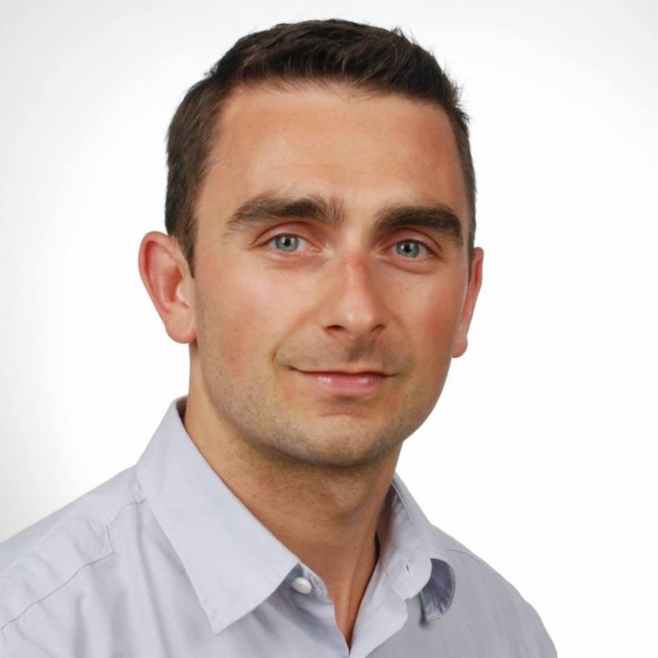 Portret mężczyzny o krótkich, ciemnych włosach i niebieskich oczach, ubranego w jasnoniebieską koszulę na białym tle. Uśmiech na twarzy, bezpośrednie spojrzenie.