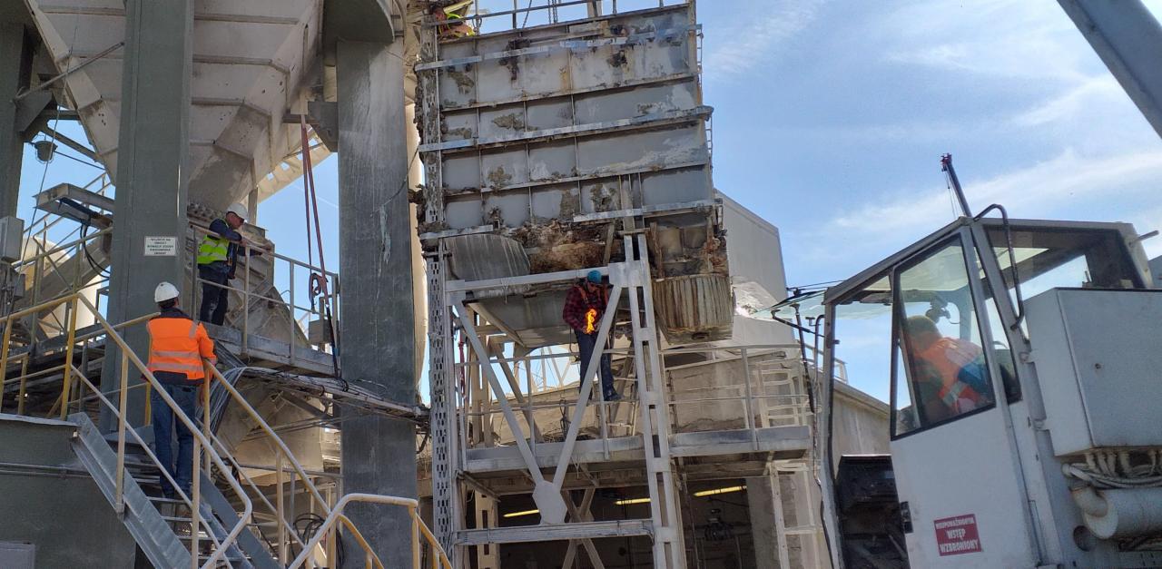 Pracownicy w kaskach i odblaskowych kamizelkach pracują przy demontażu lub naprawie konstrukcji stalowej na terenie przemysłowym, widoczny dźwig i elementy instalacji.