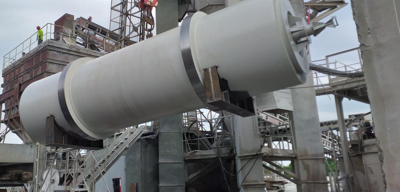 Cylindryczny element konstrukcyjny, prawdopodobnie część instalacji przemysłowej, zawieszony na dźwigu, z widocznymi stalowymi wspornikami i otoczeniem w trakcie budowy lub montażu.