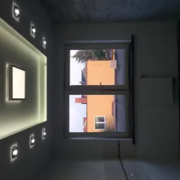 Nowoczesne oświetlenie sufitowe z wbudowanymi lampami LED i podświetleniem wnęki, widok na okno z widokiem na pomarańczowy budynek, fragment wykończonej ściany z dekoracyjną listwą.