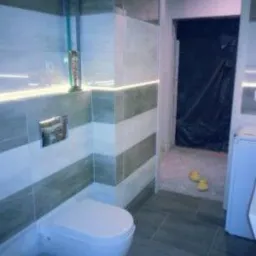 Nowoczesna łazienka z podświetlanymi płytkami w odcieniach szarości i bieli, wisząca toaleta z chromowanym przyciskiem, widoczne rury i fragment pomieszczenia w trakcie prac wykończeniowych...