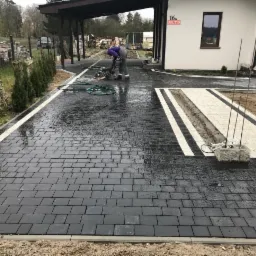 Prace brukarskie w trakcie: pracownik wyrównuje powierzchnię ciemnej kostki brukowej za pomocą maszyny, widoczne jasne pasy dekoracyjne oraz fragment domu z numerem 16A.