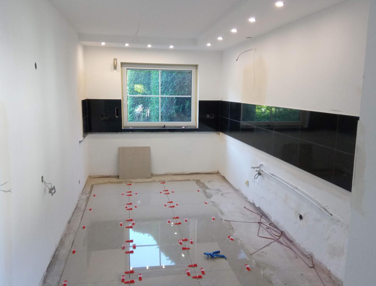 Kładzenie jasnych płytek podłogowych w kuchni z czarnymi płytkami na ścianie, widoczne krzyżyki dystansowe i poziomica.