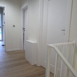 Rawel-Instal - Jasne wnętrze po remoncie: drewnopodobna podłoga, białe ściany, drzwi i schody. Minimalistyczny styl, widoczny fragment logo firmy w rogu.