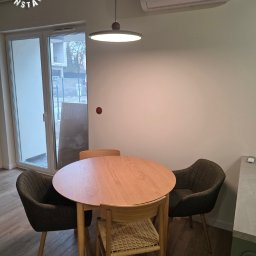 Rawel-Instal - Jasne wnętrze jadalni z okrągłym drewnianym stołem i krzesłami, oświetlone nowoczesną lampą wiszącą. Widoczny klimatyzator na ścianie i logo firmy w rogu.