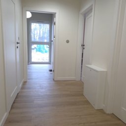 Rawel-Instal - Jasny korytarz po remoncie z panelami podłogowymi imitującymi drewno, białe ściany i drzwi. Widok na okno z zielonymi elementami na zewnątrz.