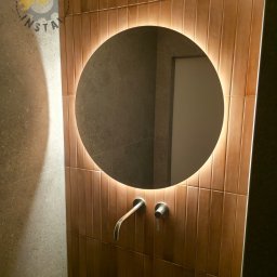 Rawel-Instal - Okrągłe lustro z podświetleniem LED nad minimalistyczną, ścienną baterią umywalkową na tle pionowo ułożonych, brązowych płytek w nowoczesnej łazience.