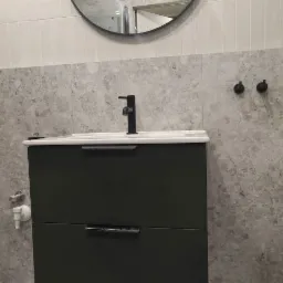 Nowoczesna łazienka po remoncie: czarna szafka pod umywalką z dwoma szufladami, biała umywalka z czarną baterią, okrągłe lustro w czarnej ramie i szare płytki imitujące marmur na ścianach i podłodze.