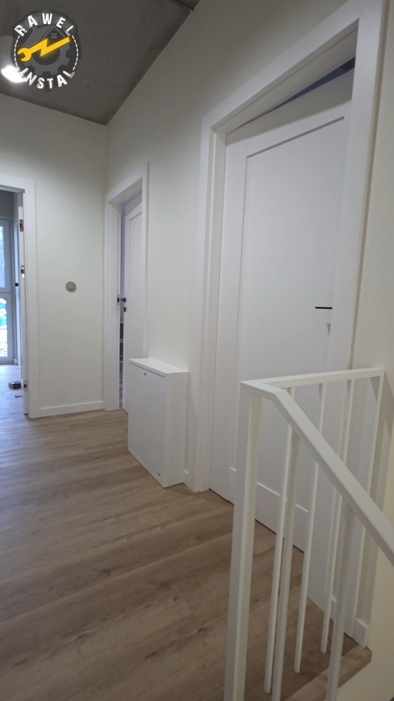 Jasne wnętrze po remoncie: drewnopodobna podłoga, białe ściany, drzwi i schody. Minimalistyczny styl, widoczny fragment logo firmy w rogu.