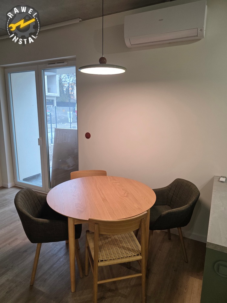 Jasne wnętrze jadalni z okrągłym drewnianym stołem i krzesłami, oświetlone nowoczesną lampą wiszącą. Widoczny klimatyzator na ścianie i logo firmy w rogu.