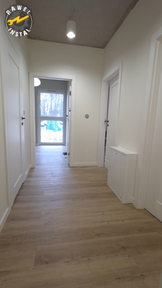 Jasny korytarz po remoncie z panelami podłogowymi imitującymi drewno, białe ściany i drzwi. Widok na okno z zielonymi elementami na zewnątrz.