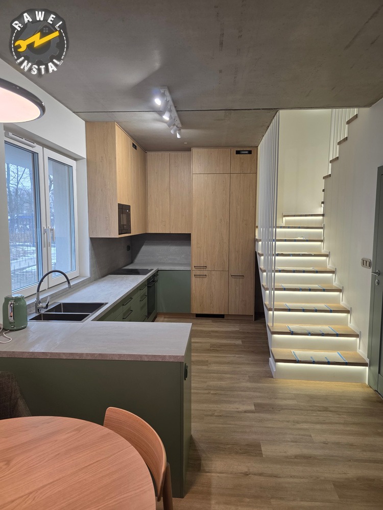 Nowoczesna kuchnia z drewnianymi szafkami, zielonymi akcentami i oświetlonymi schodami. Minimalistyczny design, jasne wnętrze, widok na okno i stół z krzesłami.