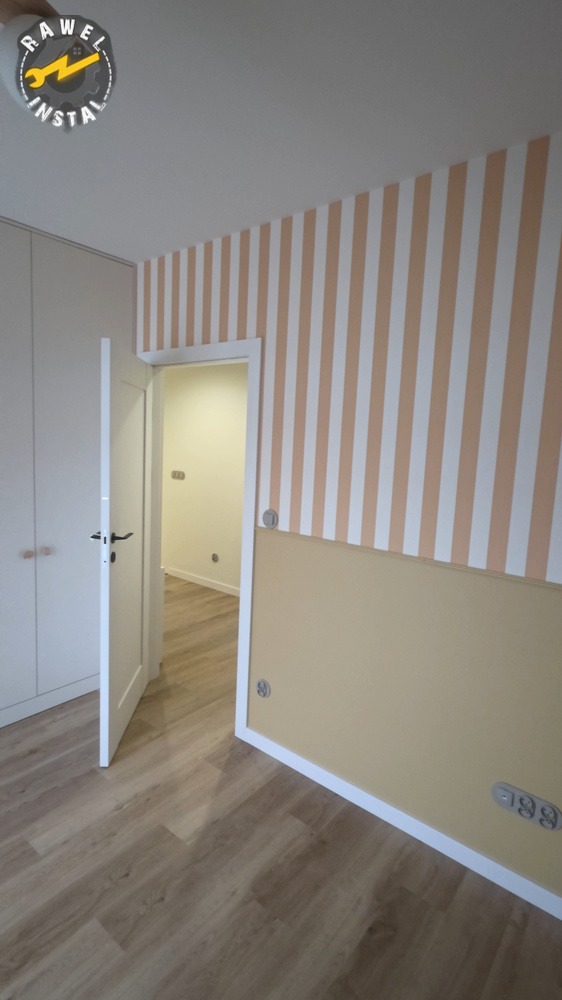 Pokój po remoncie: ściany w pasy beżowo-białe, dolna część beżowa, drewniana podłoga, białe drzwi otwarte, widok na kolejne pomieszczenie.