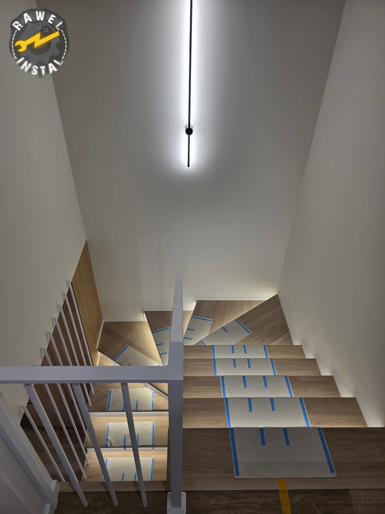 Nowoczesne schody z drewnianymi stopniami, zabezpieczone folią i taśmą malarską, oświetlone listwą LED na suficie. Widok z góry, białe barierki.