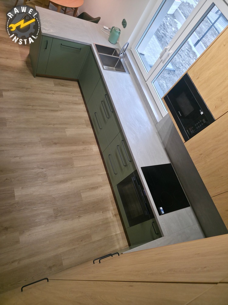 Nowoczesna kuchnia w odcieniach zieleni i drewna z blatem imitującym beton, z widoczną płytą indukcyjną i piekarnikiem. Ujęcie z góry, prezentujące układ i wykończenie.