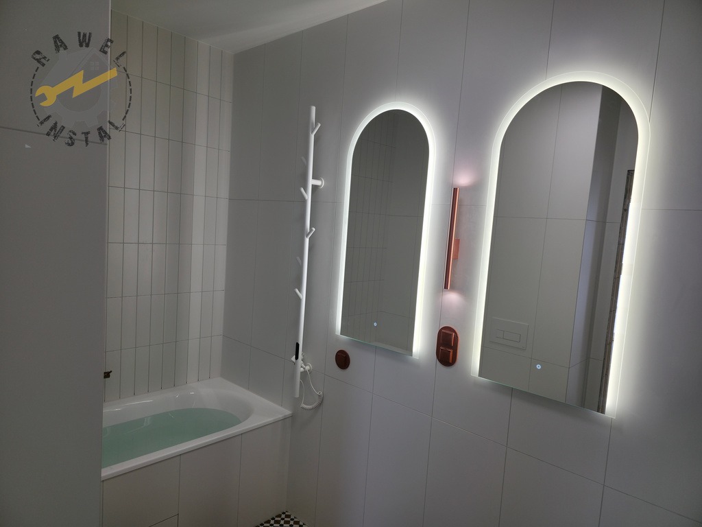 Biała łazienka z wanną, dwoma podświetlanymi lustrami w kształcie łuku i minimalistycznym wieszakiem na ręczniki. Nowoczesny design, jasne płytki.