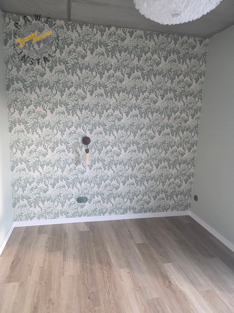 Pokój po remoncie: wzorzysta tapeta w odcieniach zieleni, panele podłogowe imitujące drewno, biała listwa przypodłogowa. Widoczne gniazdka elektryczne i lampa sufitowa.