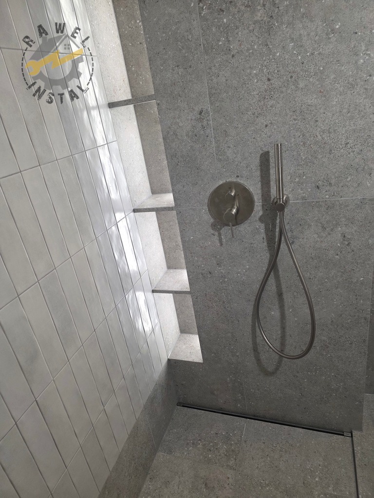 Wykończona kabina prysznicowa z szarymi płytkami imitującymi kamień i białymi, wąskimi płytkami na ścianie. Widoczne półki wnękowe i bateria prysznicowa.