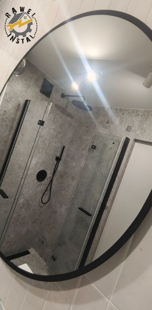 Owalne lustro z czarną ramą odbija wnętrze wyremontowanej łazienki z kabiną prysznicową ze szklanymi drzwiami, deszczownicą i panelami ściennymi imitującymi szary kamień.