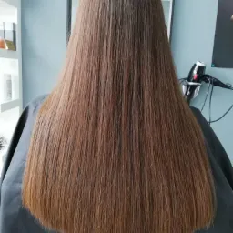 Długie, proste, brązowe włosy po zabiegu prostowania, widoczne od tyłu, z suszarką do włosów w tle i peleryną ochronną.