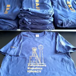 Niebieska koszulka z nadrukiem przedstawiającym szyb kopalni Ignacy w Rybniku, leżąca na tle ułożonych w stosy innych koszulek z napisem 'Raj'