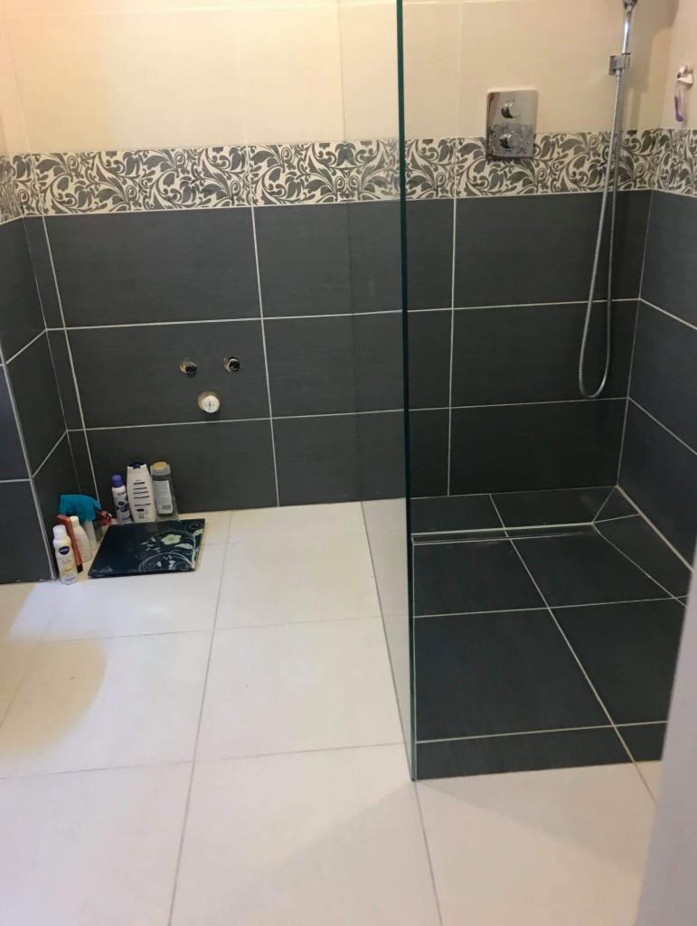 Nowoczesna kabina prysznicowa z szarymi płytkami, ozdobnym pasem dekoracyjnym i białymi płytkami podłogowymi, widoczne kosmetyki na podłodze.