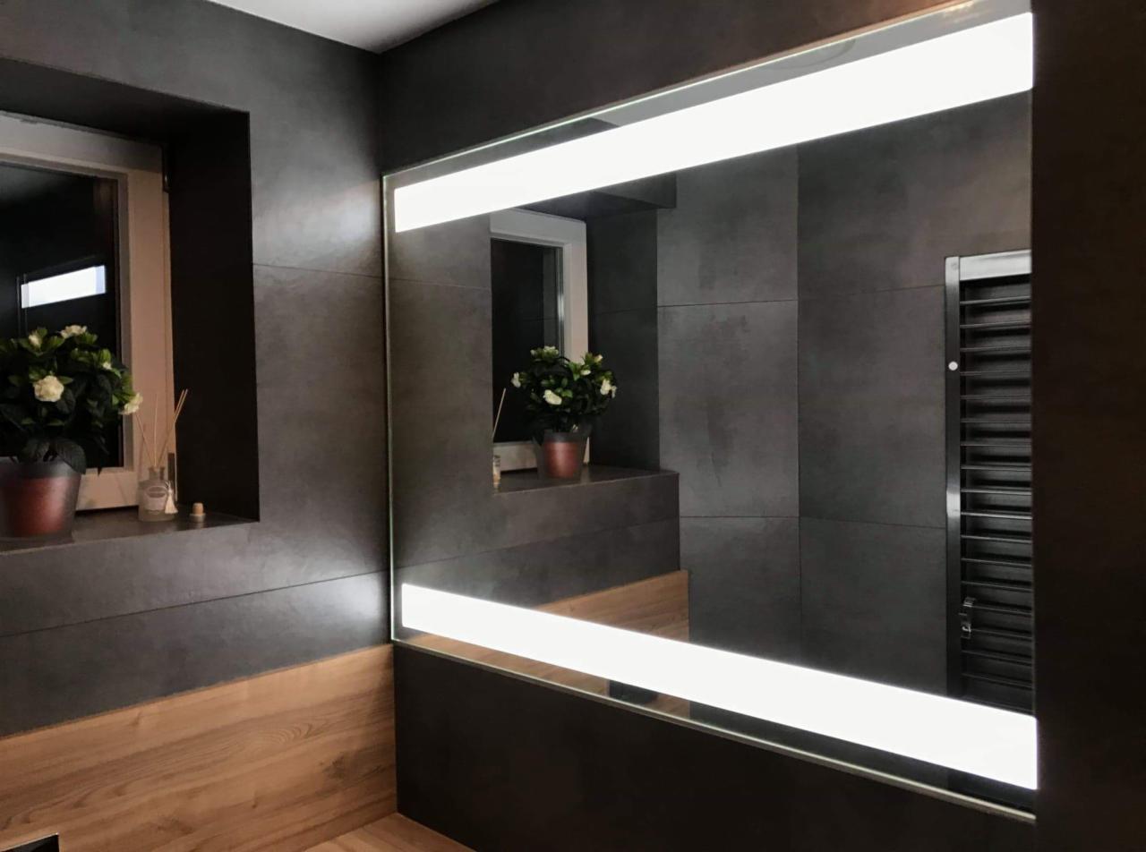 Nowoczesna łazienka z szarymi płytkami, podświetlanym lustrem i drewnianymi panelami, odbicie okna z kwiatami w lustrze.