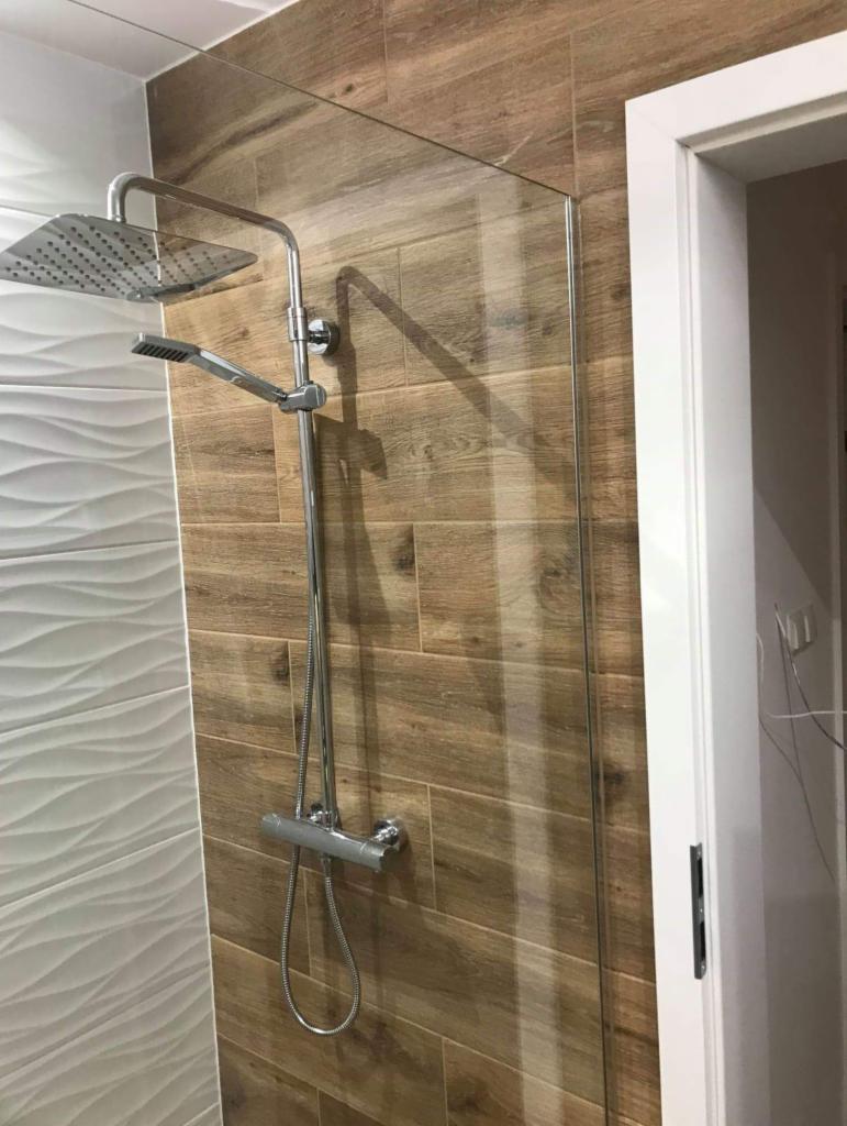 Nowoczesna kabina prysznicowa typu walk-in w Warszawie, z chromowaną armaturą i deszczownicą, na ścianach płytki imitujące drewno oraz białe, faliste płytki.