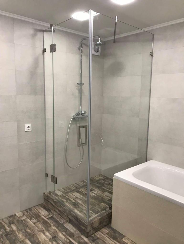 Szklana kabina prysznicowa z deszczownicą i baterią, zamontowana na podwyższeniu z płytek imitujących drewno, obok wanna w jasnej obudowie, ściany wyłożone szarą płytką.