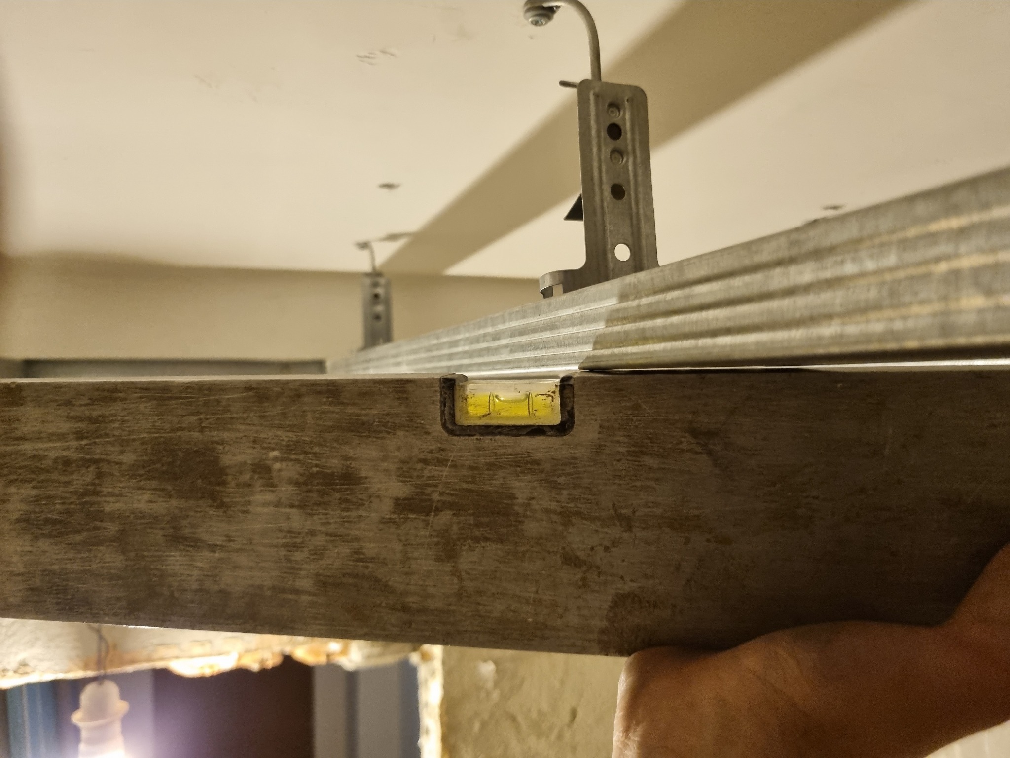 Poziomica przykładana do metalowego stelażu podwieszanego sufitu, widoczne uchwyty mocujące do stropu.