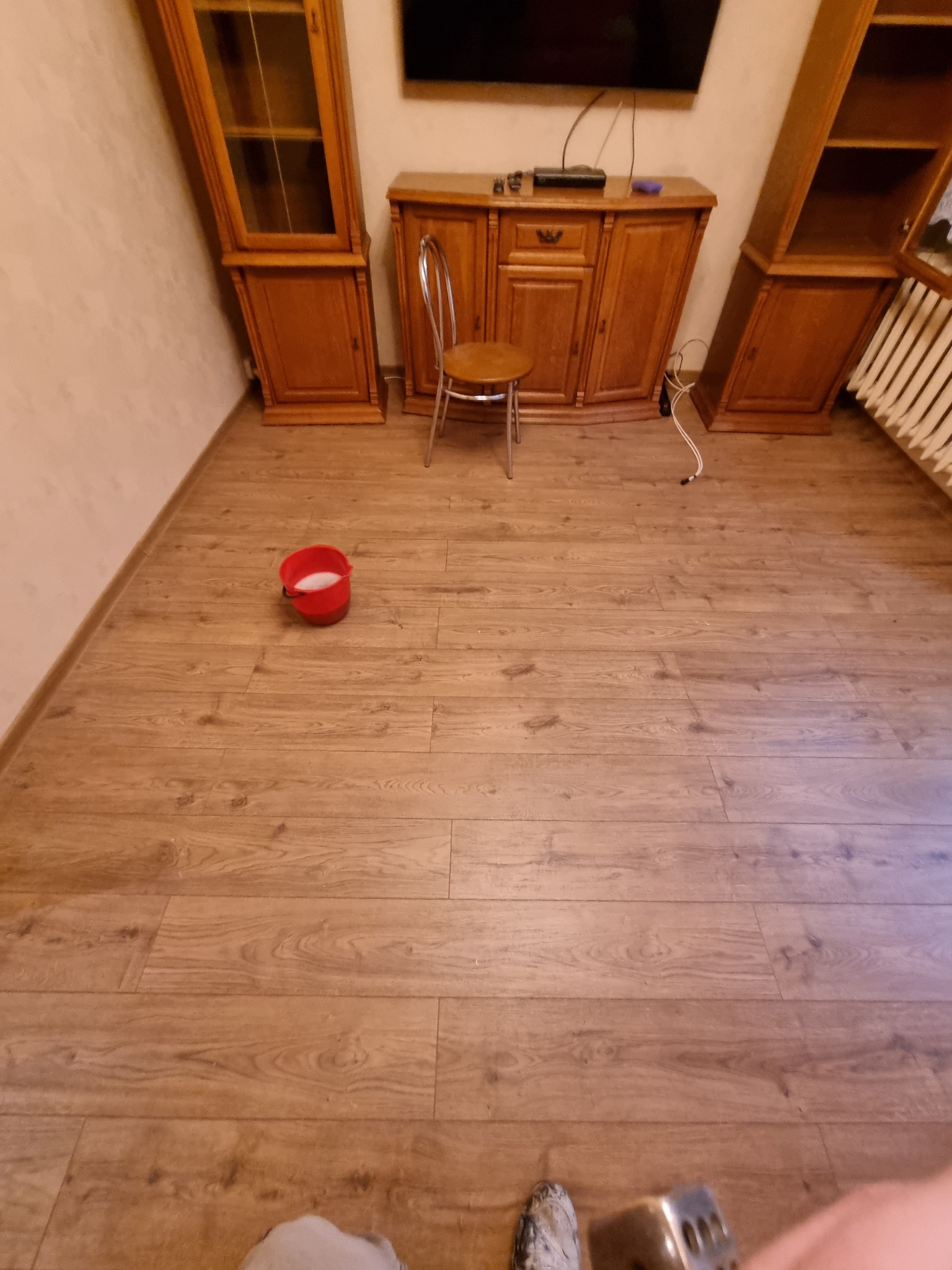 Pokój z drewnianą podłogą, wiadro z białą farbą i meblami w tle, przygotowany do prac wykończeniowych.