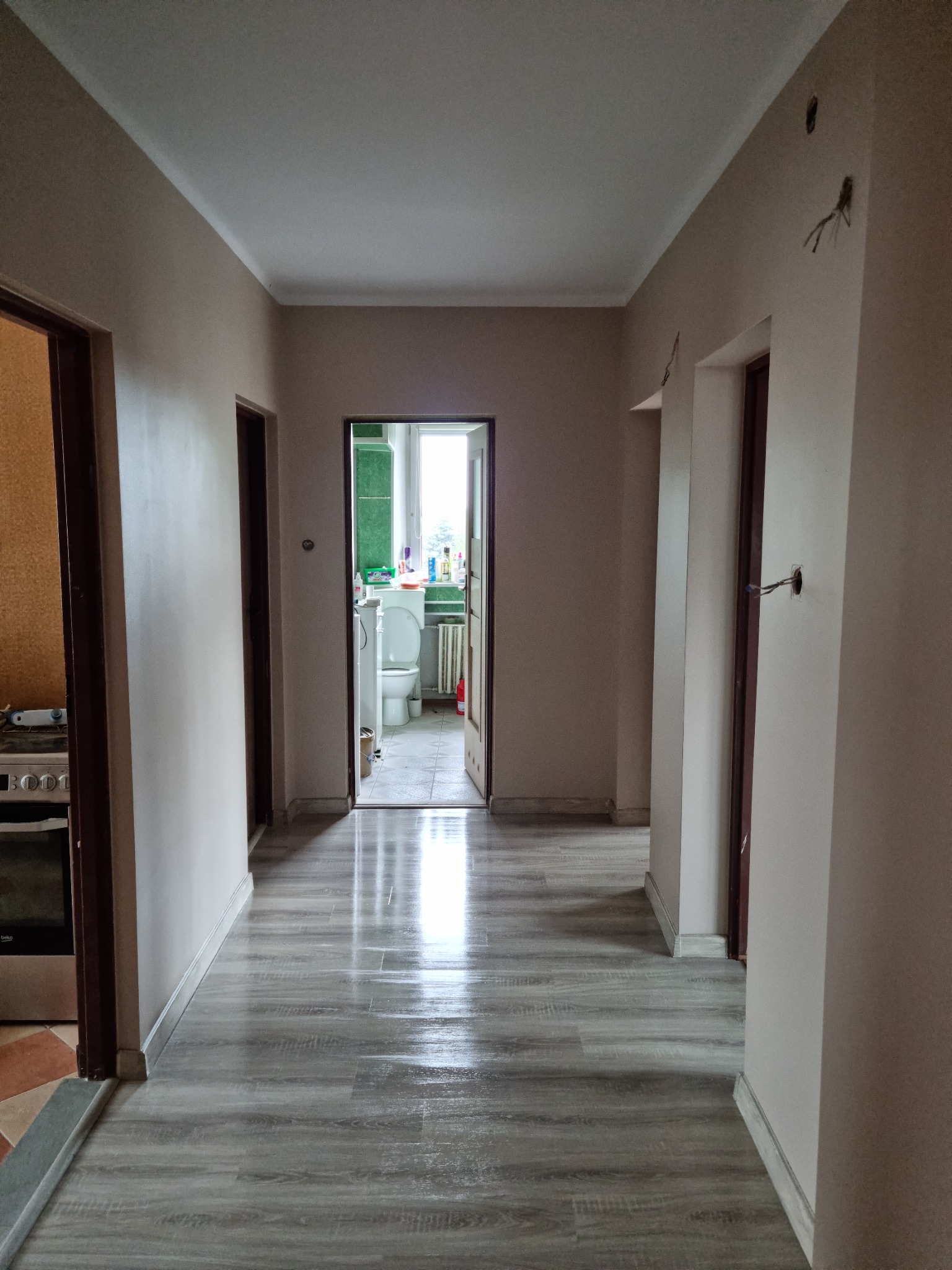 Korytarz w mieszkaniu po remoncie, widoczne świeżo położone panele podłogowe w odcieniach szarości imitujące drewno, ściany pomalowane na jasny beż, widoczne otwarte drzwi do kuchni i łazienki...