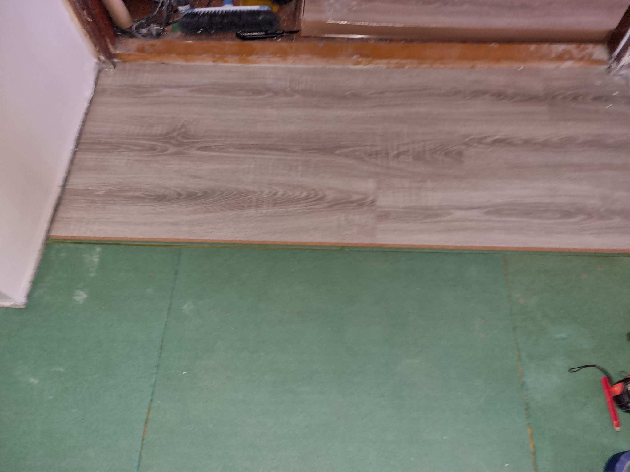 Fragment podłogi z ułożonymi panelami imitującymi drewno, widoczne zielone podłoże pod panele, przy ścianie widoczne niedociągnięcia w wykończeniu.