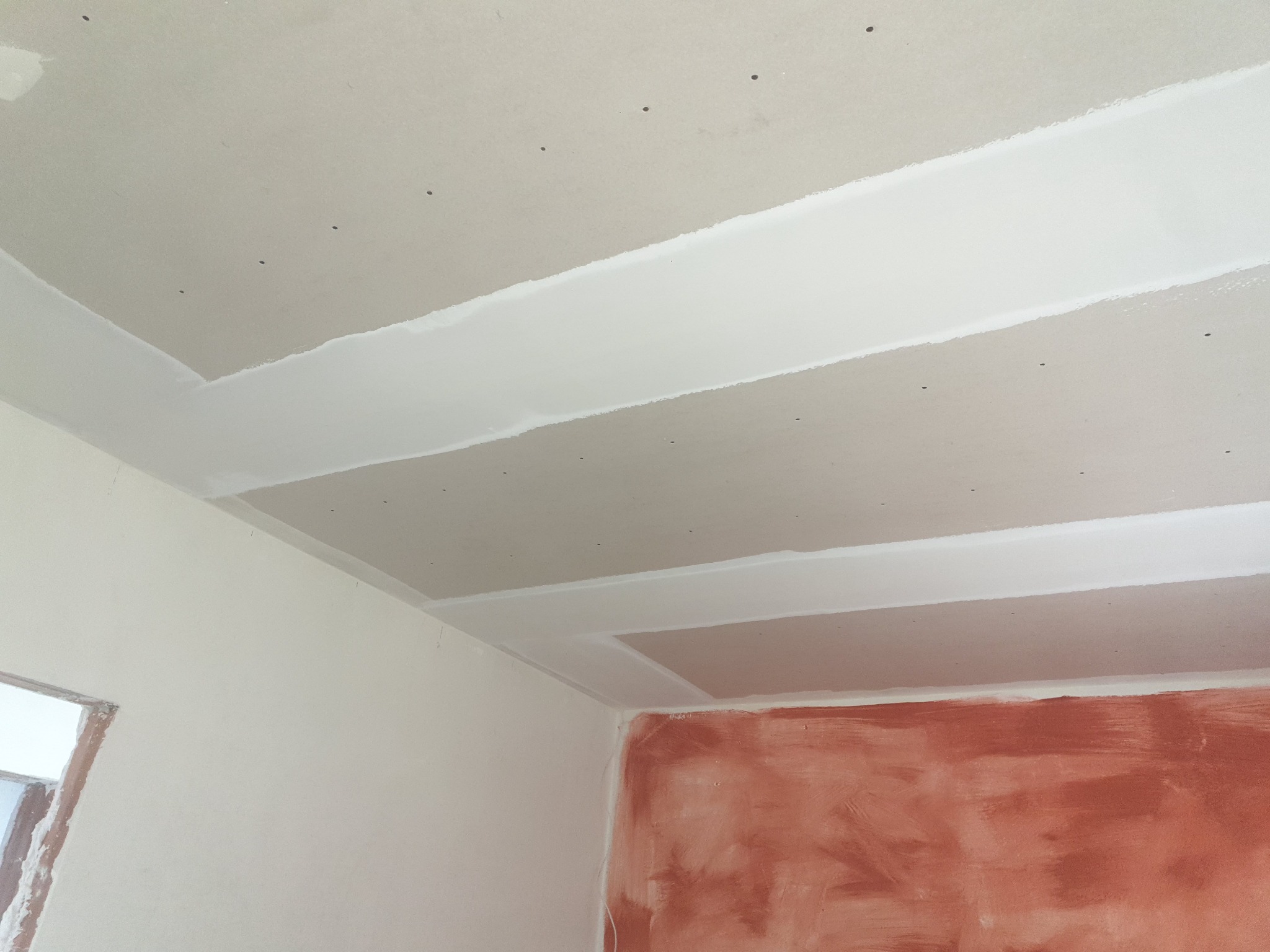 Zamontowany sufit z płyt kartonowo-gipsowych ze śladami po wkrętach, z widocznymi łączeniami płyt wypełnionymi masą szpachlową, na tle fragmentu ściany z widocznymi śladami po malowaniu.