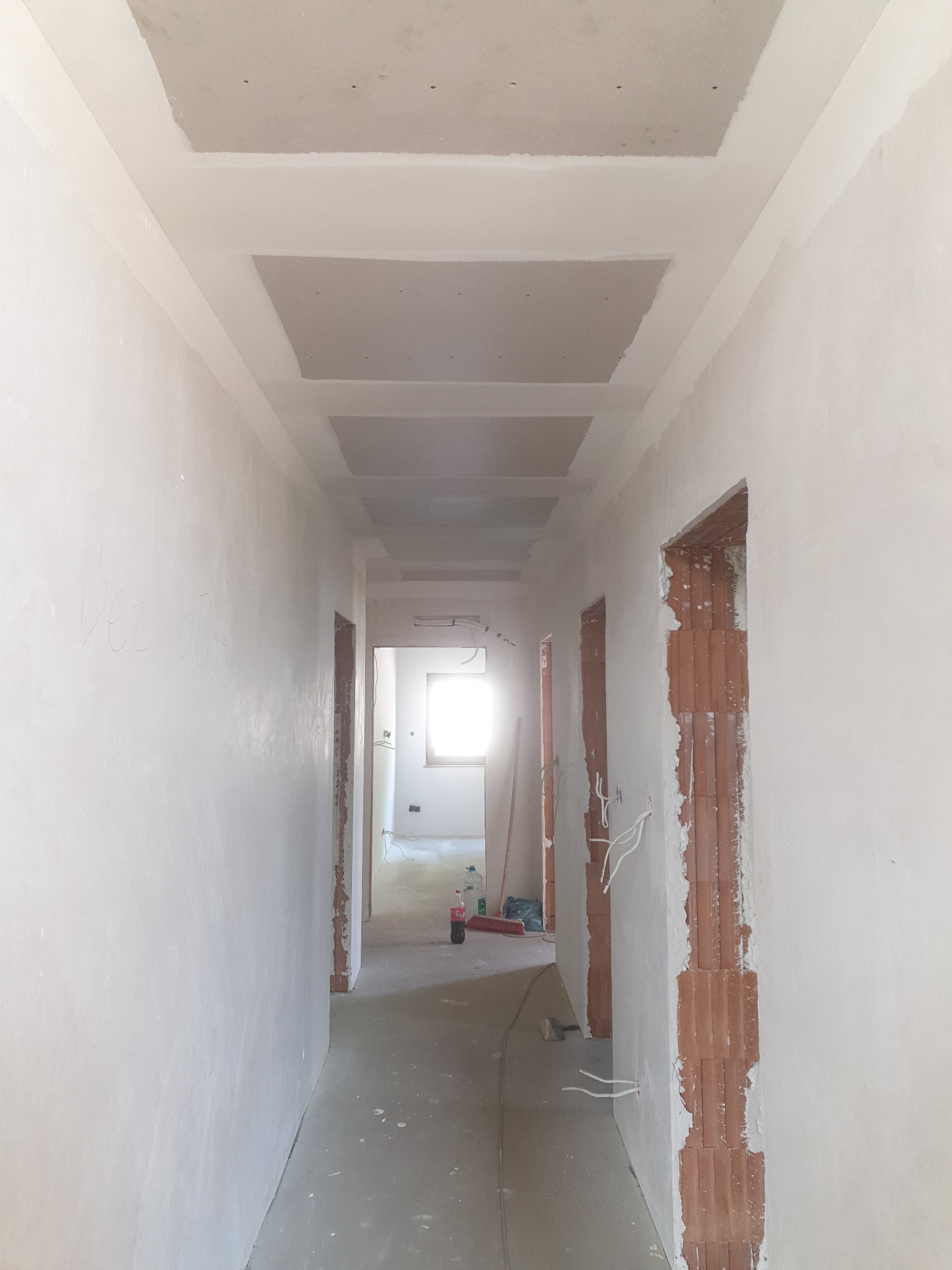 Korytarz w trakcie prac przygotowawczych do wykończenia, widoczne płyty kartonowo-gipsowe na suficie, ściany po szpachlowaniu, niezabezpieczone otwory drzwiowe i wystające przewody elektryczne.