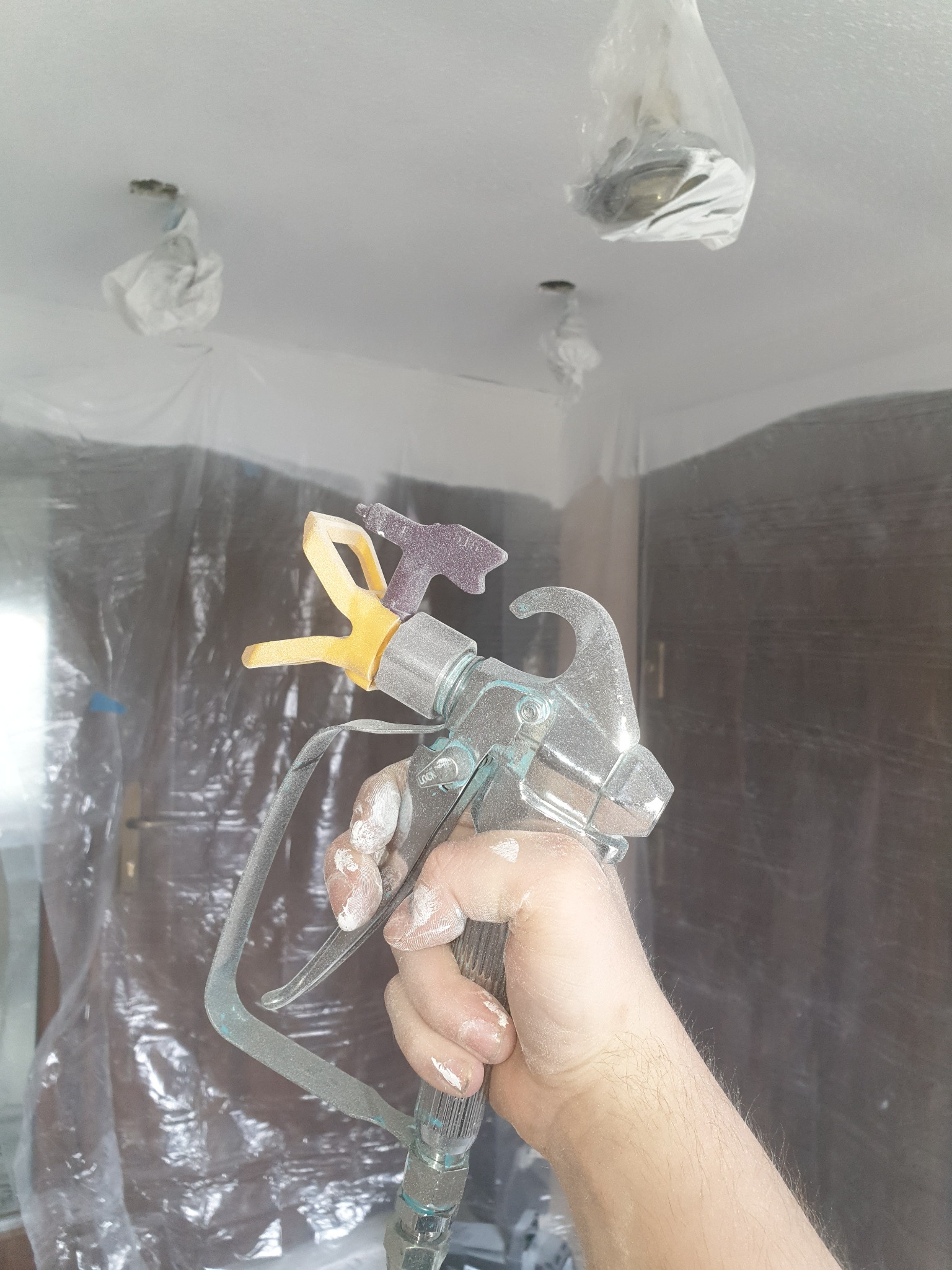 Pistolet natryskowy w dłoni pracownika, widoczne ślady farby na dłoni, w tle zabezpieczone folią pomieszczenie przed malowaniem, lampy owinięte folią ochronną.