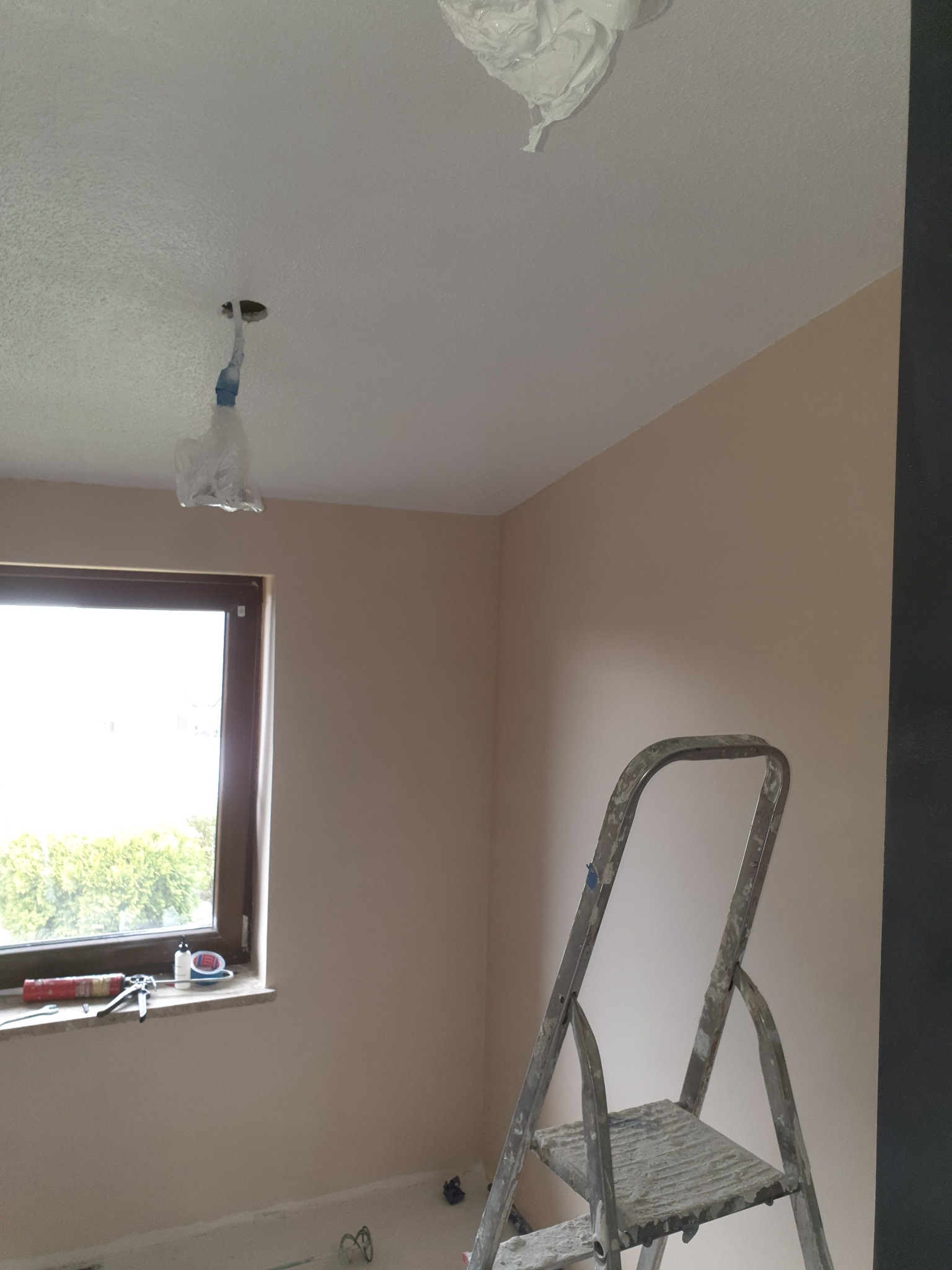 Pokój w trakcie odświeżania ścian w odcieniach beżu, z zabezpieczonymi folią lampami sufitowymi, drabina malarska z widocznymi śladami farby oraz narzędzia przy oknie.