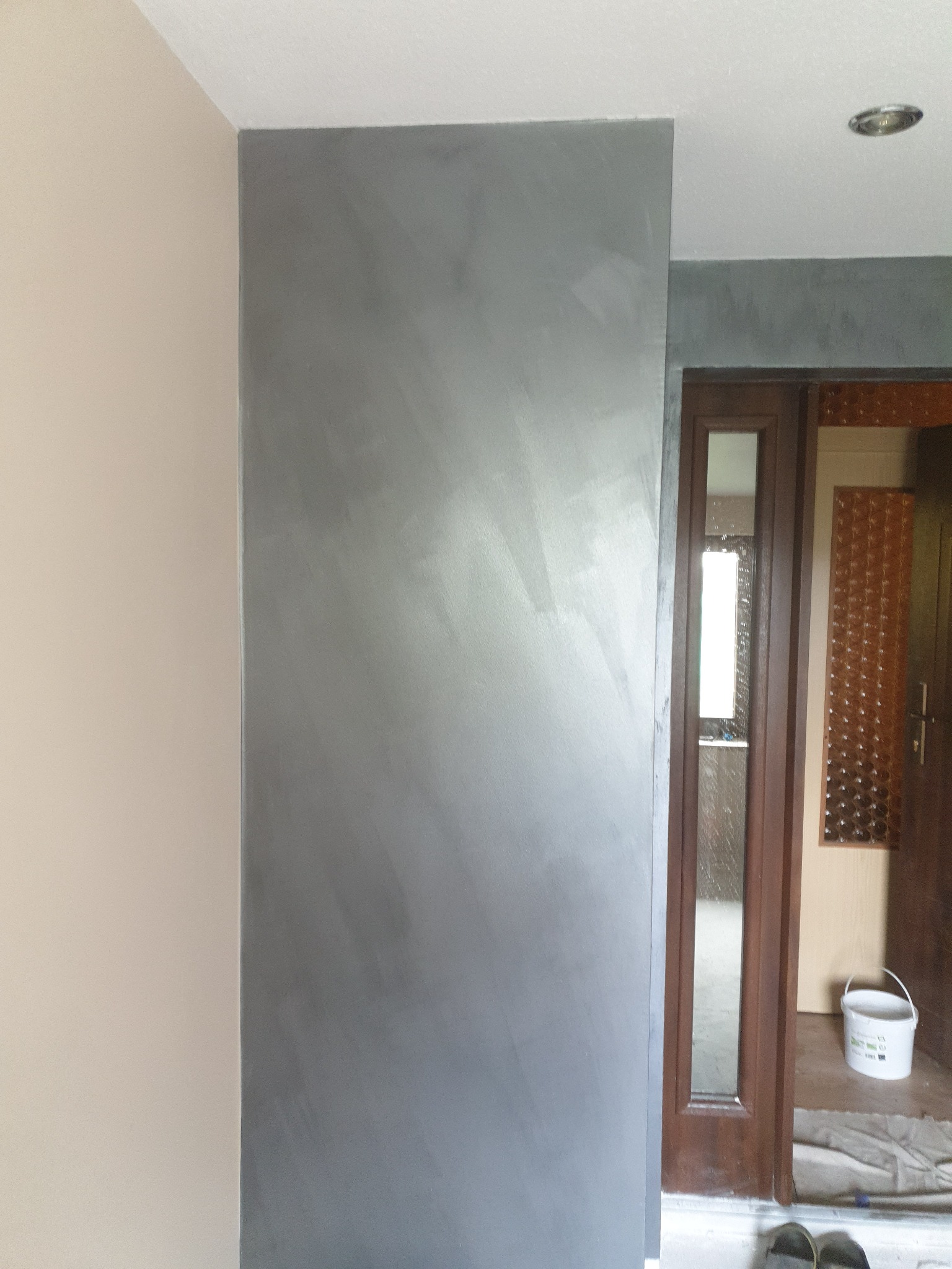 Ściana po wykonaniu gładzi dekoracyjnej w odcieniach szarości, widoczny fragment sąsiedniej ściany w kolorze beżowym i drzwi z dekoracyjną szybą oraz wiadro z farbą w tle.