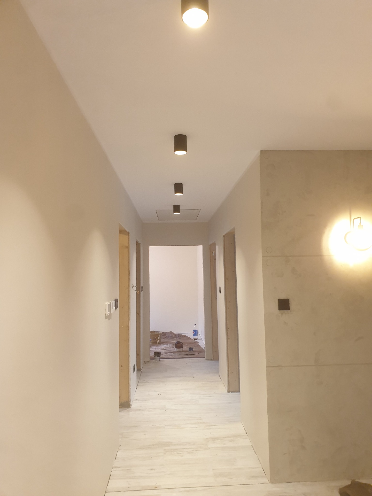 Korytarz w trakcie prac wykończeniowych: ściany przygotowane pod malowanie, widoczne instalacje elektryczne, podłoga częściowo zabezpieczona, sufit z zamontowanymi punktowymi lampami.