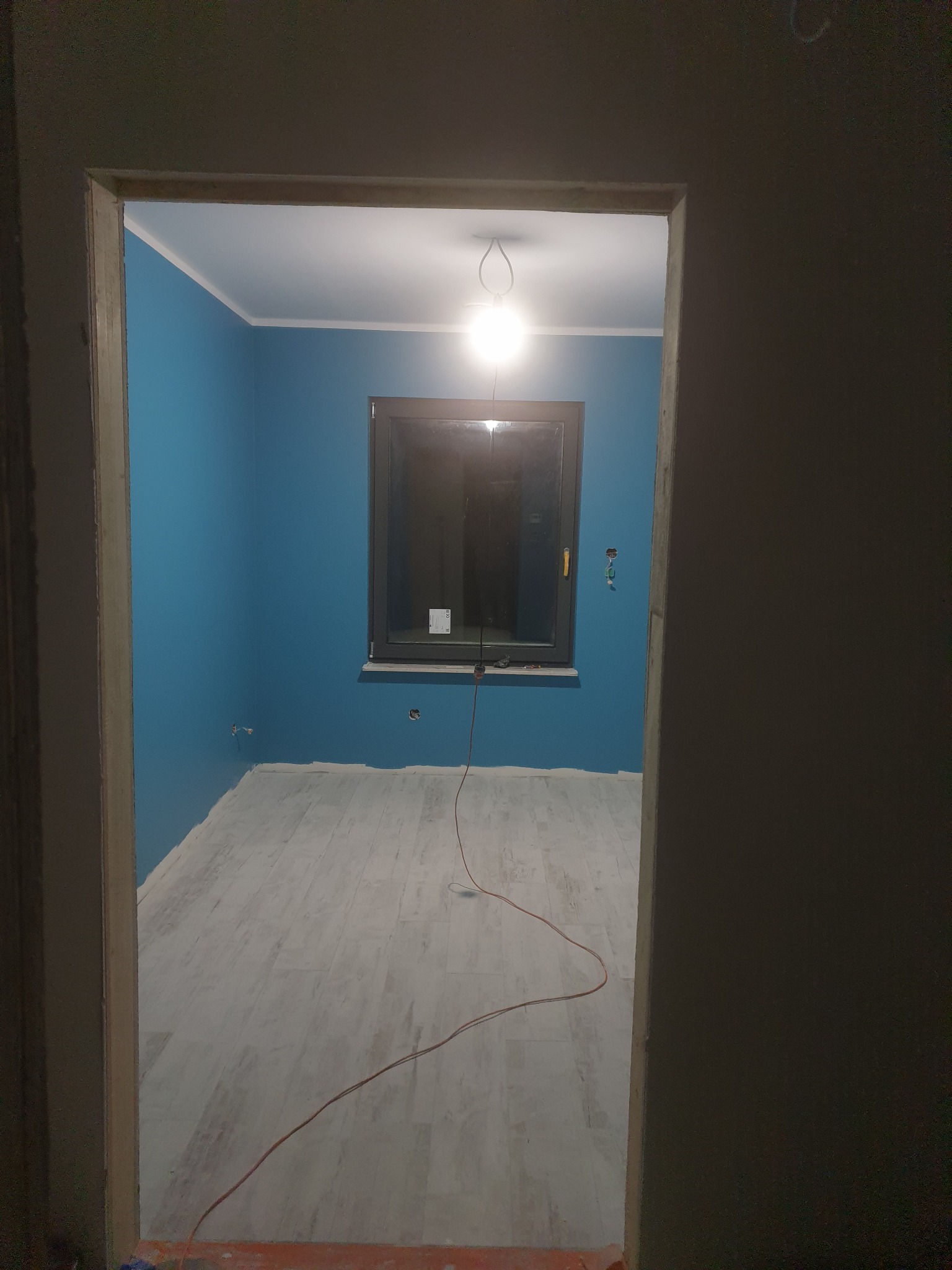 Pokój po remoncie z pomalowanymi na niebiesko ścianami, jasną podłogą i oknem, widok z korytarza przez surową ościeżnicę drzwiową.