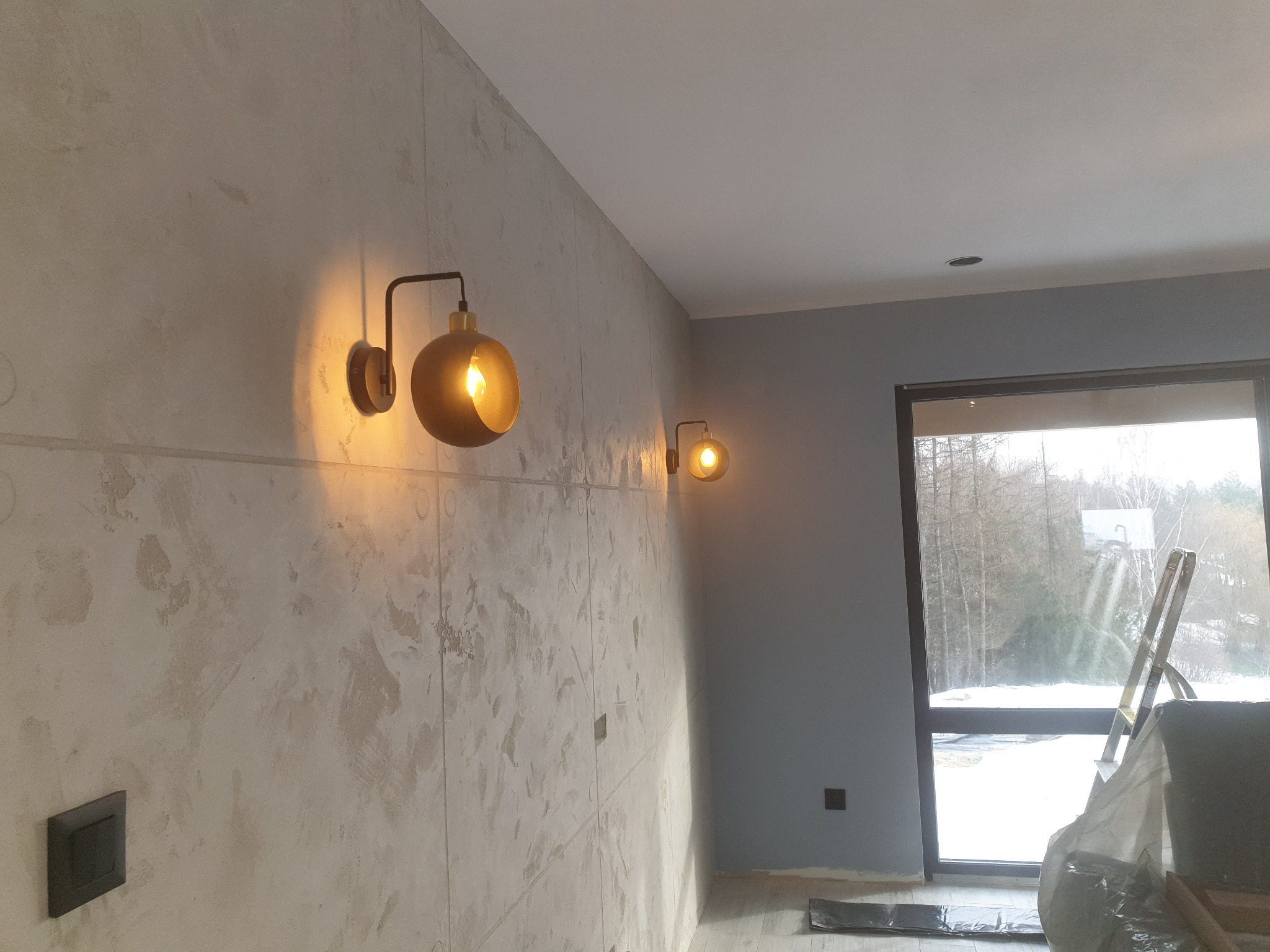 Ściana z dekoracyjną fakturą imitującą beton, oświetlona dwoma złotymi kinkietami, widoczne gniazdko elektryczne i fragment okna z drabiną w tle.